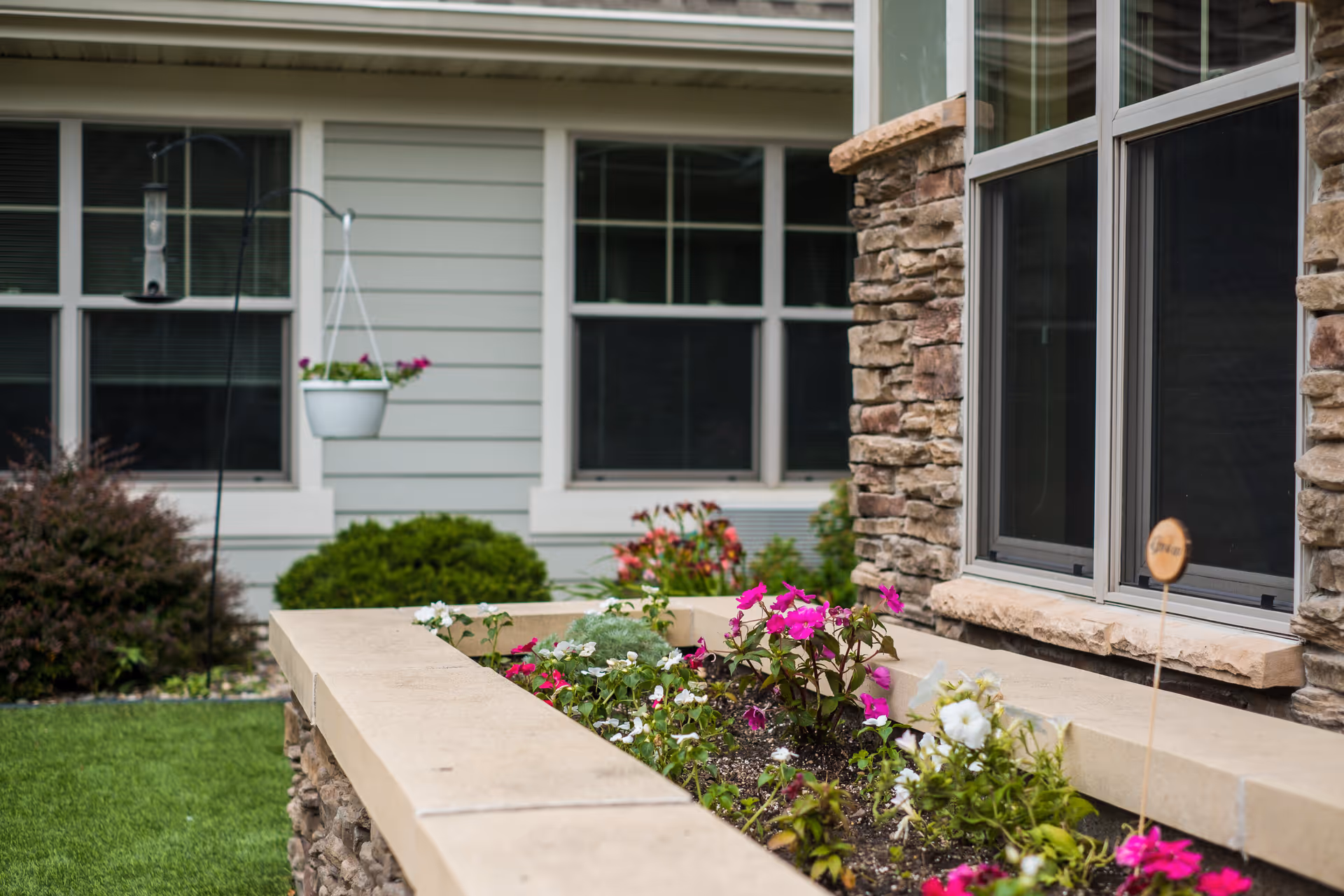 A close-up view of a flower bed with colorful flowers in front of a building with stone and siding exterior walls and windows. There is a hanging white flower pot and a bird feeder in the background, along with green bushes and grass.