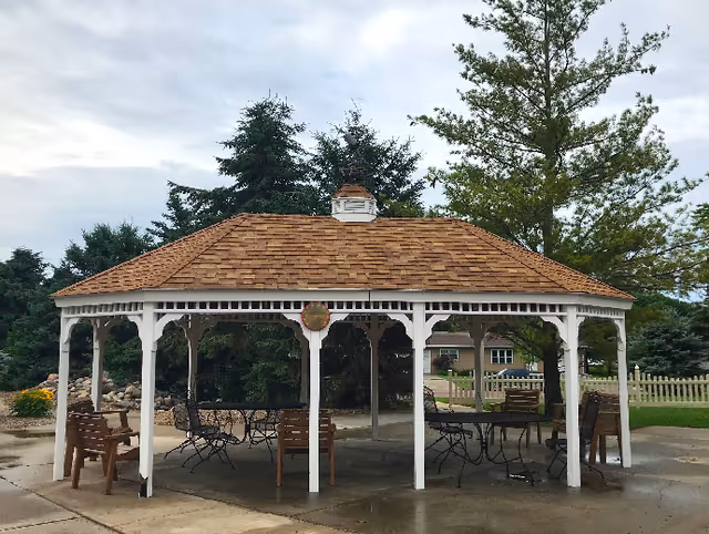 A white wooden gazebo with a shingled roof covering outdoor tables and chairs on a paved area, surrounded by trees and lawn.