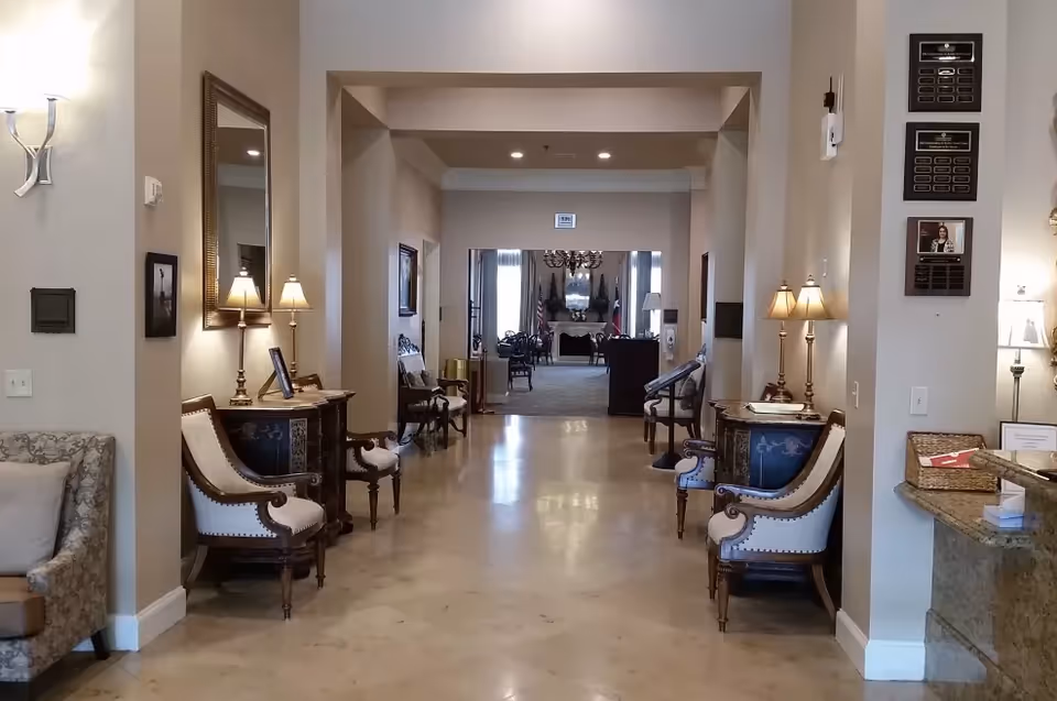 Interior hallway of a senior living facility with polished floors, antique-style chairs and side tables with lamps on both sides. The hallway leads to a room with a fireplace, chandelier, and flags visible in the background.