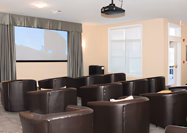 A room with multiple dark brown leather armchairs arranged in rows facing a large screen on the wall, with beige walls, a window with curtains, and a projector mounted on the ceiling.
