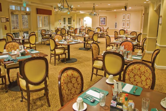 A spacious dining room with multiple wooden tables and upholstered chairs arranged neatly. Each table is set with placemats, napkins, utensils, cups, and condiments. The room has warm lighting, chandeliers, framed artwork on the walls, and large windows with curtains.