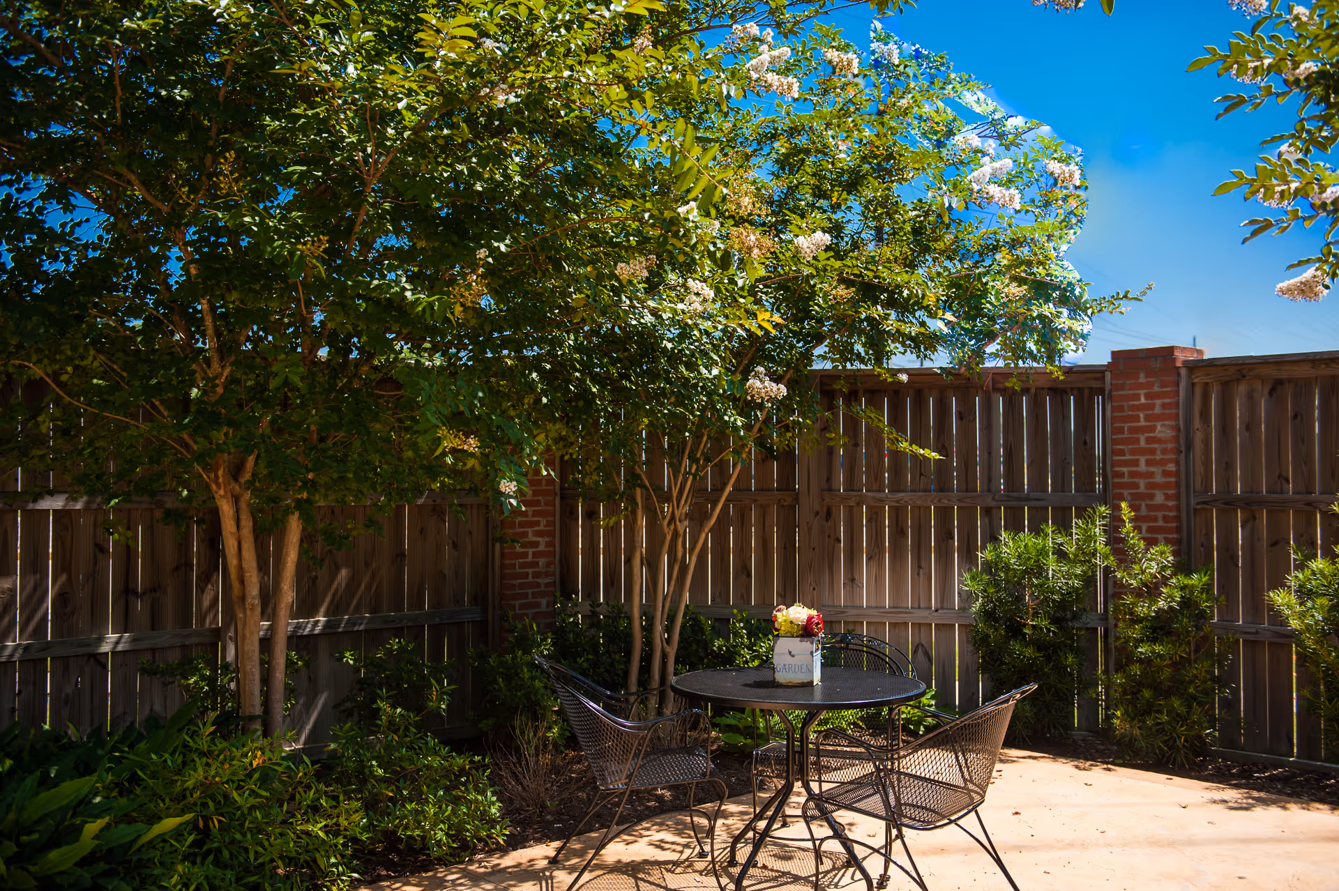 Shaded outdoor patio with a round metal table and chairs beneath trees beside a wooden fence.