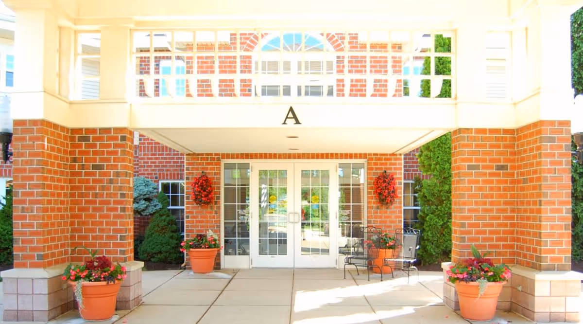 Entrance to a building with brick pillars and white framed glass double doors. There are large potted plants with flowers on either side of the entrance and hanging flower baskets on the brick walls. The letter 'A' is displayed above the doorway.