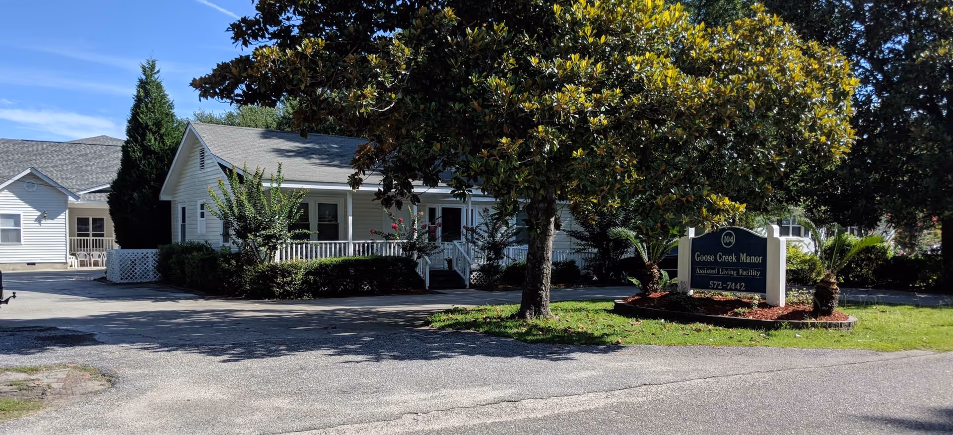 Exterior view of Goose Creek Manor, an assisted living facility, showing a white building with a porch surrounded by trees and shrubs. A sign in front reads 'Goose Creek Manor Assisted Living Facility' with a phone number. The sky is clear and blue.