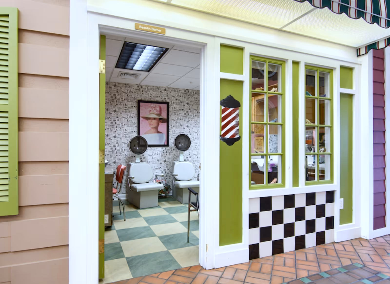Interior view of a beauty/barber room in a senior living facility. The room has two white salon chairs with hair dryers above them, a framed picture of a woman wearing a hat on the wall, and a checkered black and white pattern on the lower part of the exterior wall. The door frame and window frames are painted white and green, with a barber pole decoration on the wall next to the window.