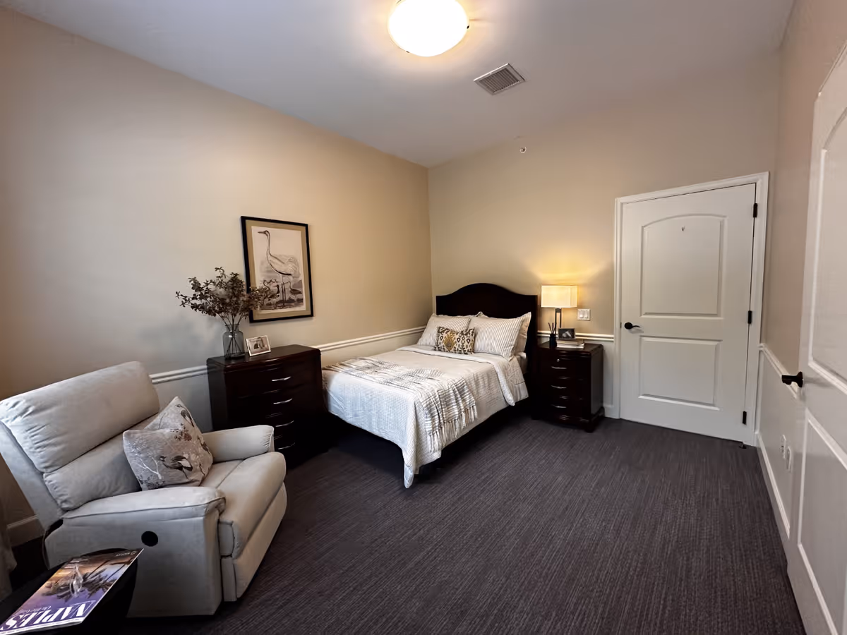 A cozy bedroom with a single bed featuring white and beige bedding, a dark wooden headboard, and matching nightstands on either side. One nightstand has a lamp and a small picture frame, while the other holds a vase with dried flowers and a framed picture. There is a comfortable beige recliner chair with a decorative pillow next to a small side table with a magazine. The walls are painted light beige, and there is a framed picture of a bird hanging above the nightstand. The room has a carpeted floor and a closed white door.