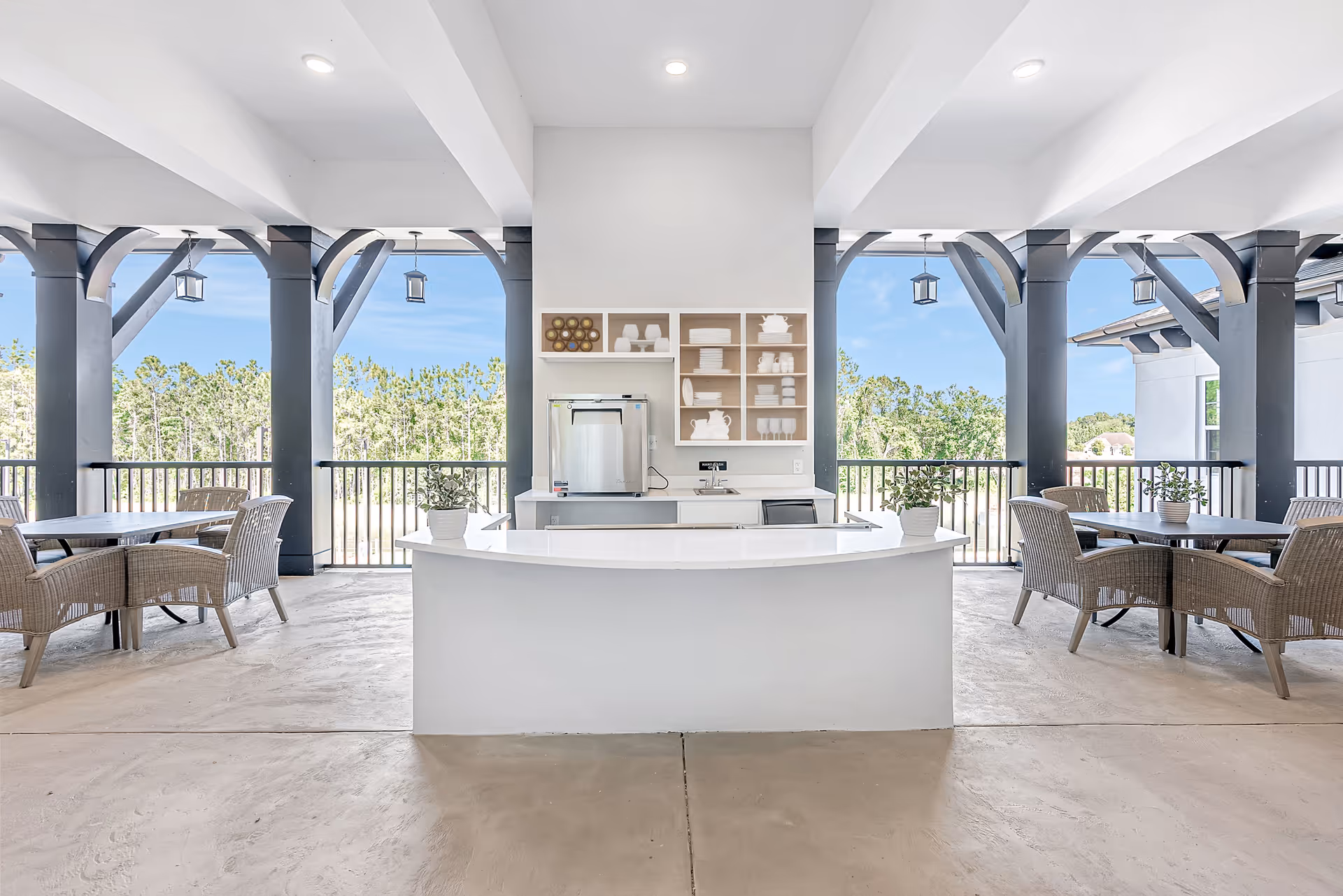 Outdoor covered seating area with a white curved counter in the center, wicker chairs around tables on either side, black structural beams, and a view of trees and blue sky in the background.