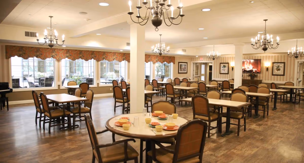 A spacious dining room with multiple tables and chairs arranged neatly. The tables are set with plates, bowls, cups, and utensils. Large windows with floral valances allow natural light to fill the room. Chandeliers hang from the ceiling, and a fireplace with a decorative mantel is visible on the far wall.