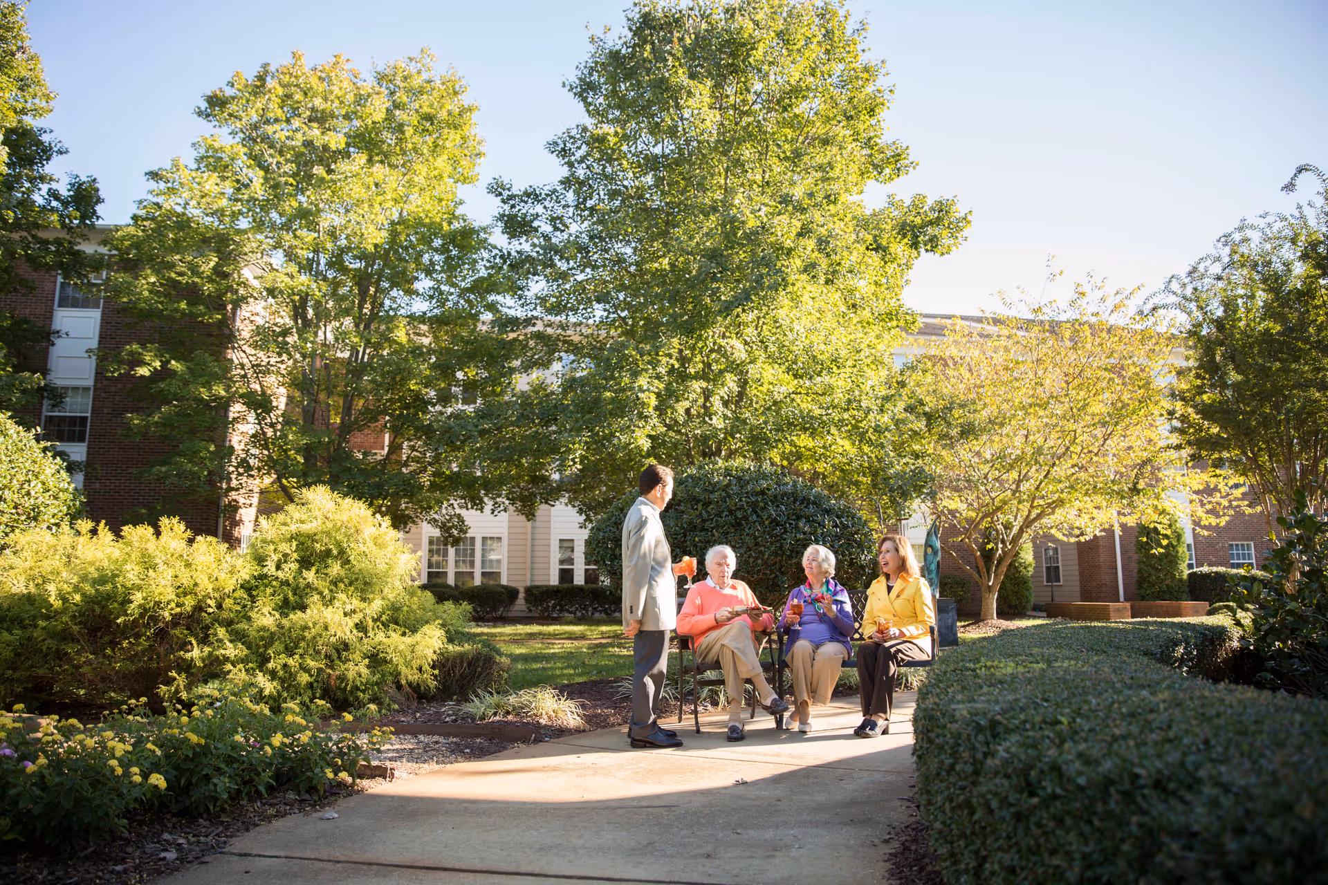 Three elderly women sitting on chairs outdoors in a garden area, holding drinks and talking, with a man standing nearby. They are surrounded by green trees and bushes, with a building visible in the background under a clear sky.