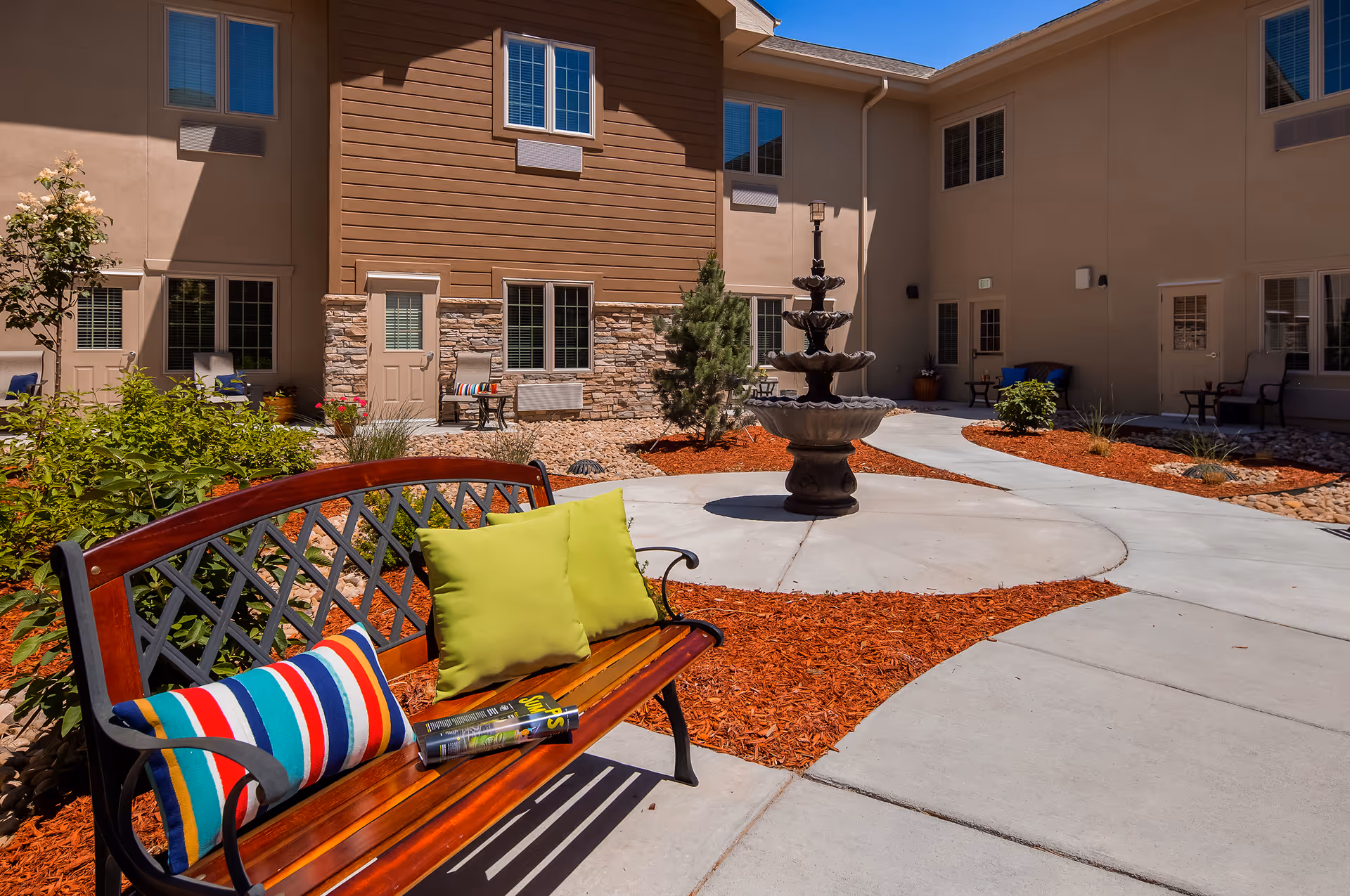 Outdoor courtyard area of a senior living facility with a wooden bench adorned with colorful cushions and a magazine, a multi-tiered water fountain, landscaped garden beds with red mulch, and beige building walls with windows and doors surrounding the courtyard.