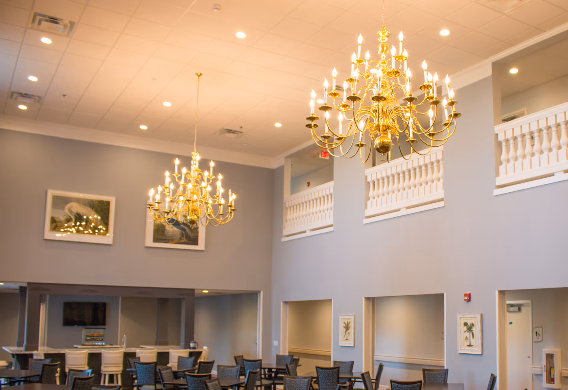 A spacious dining area with multiple tables and chairs, two large ornate gold chandeliers hanging from a high ceiling, light gray walls with framed artwork, and a balcony with white railings overlooking the room.