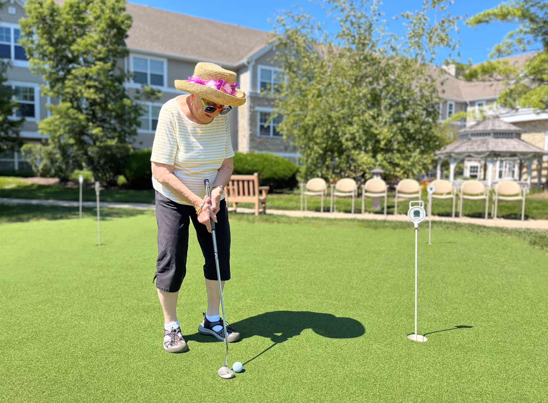 An elderly woman wearing a straw hat with pink decorations, sunglasses, a striped shirt, and black pants is playing mini golf on a putting green outside. Behind her are chairs, a wooden bench, trees, and a building with multiple windows under a clear blue sky.