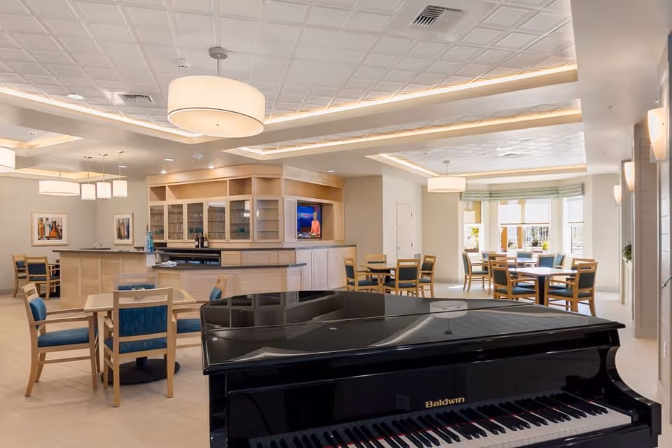 A spacious and well-lit common area featuring a black Baldwin grand piano in the foreground, multiple wooden tables with blue cushioned chairs, a bar area with glass cabinets and a TV, and large windows allowing natural light to fill the room.