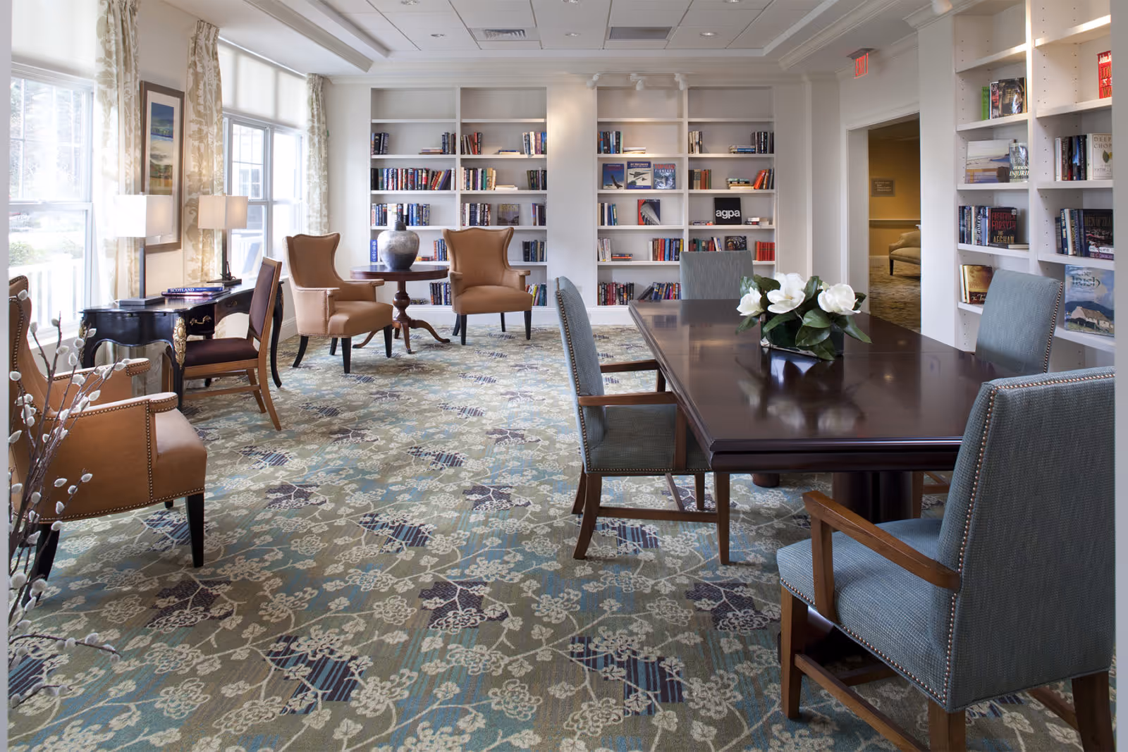 A bright and spacious library room with large windows letting in natural light. The room features built-in white bookshelves filled with books, a dark wooden table with four upholstered chairs, and two brown armchairs around a small round table. The carpet has a floral pattern in shades of blue, green, and beige. There are decorative elements including a vase on the table and framed artwork on the wall.