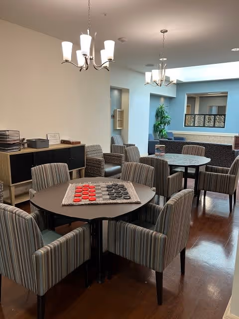 A cozy common area with round tables and striped upholstered chairs arranged around them. One table has a checkers board with red and black pieces set up. The room has wooden flooring, modern chandeliers, and a sideboard with office supplies. In the background, there is a seating area with a plant and a window opening to another room.