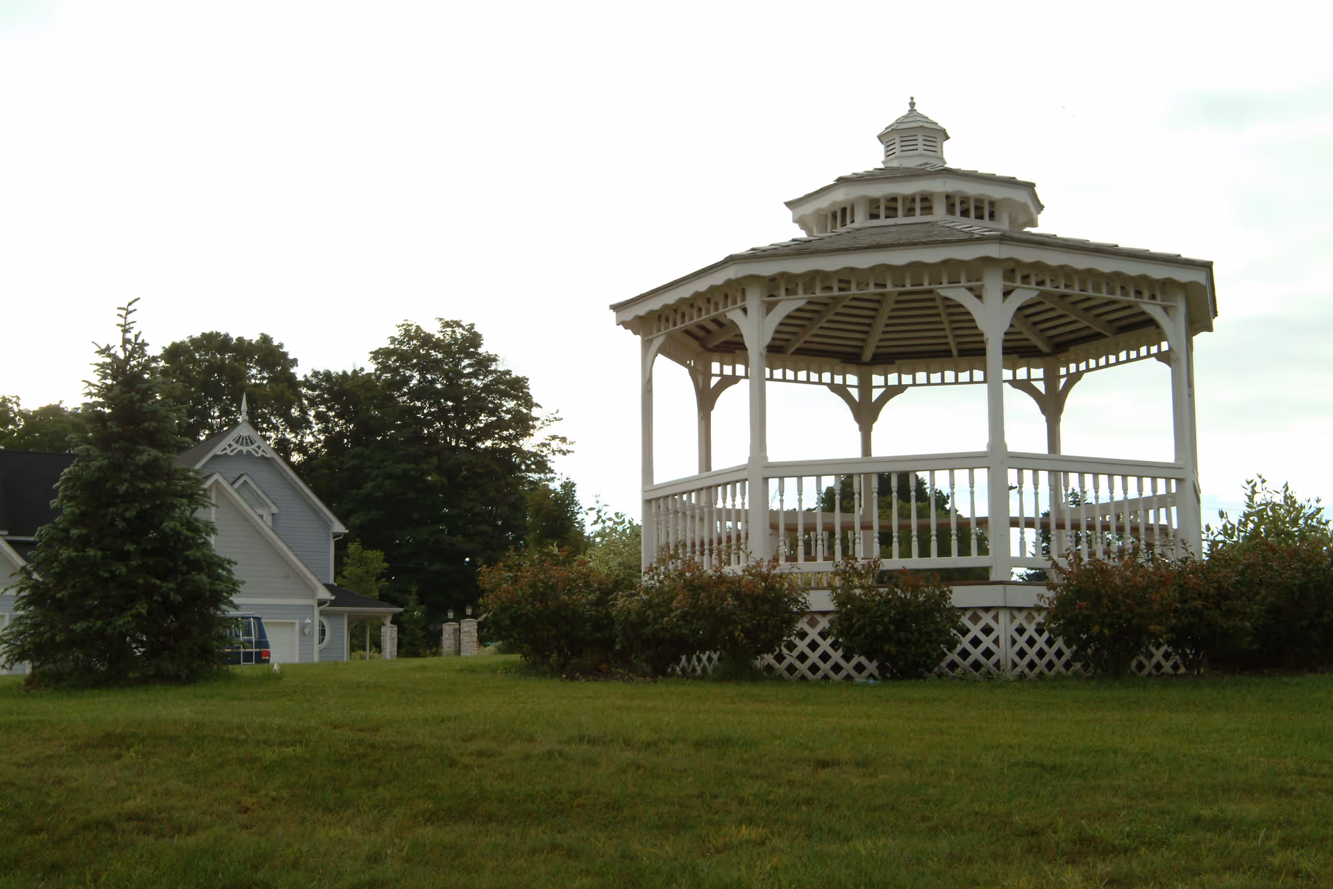 A white wooden gazebo with a decorative roof sits on a grassy lawn surrounded by bushes. In the background, there is a gray house partially visible with trees behind it under a cloudy sky.
