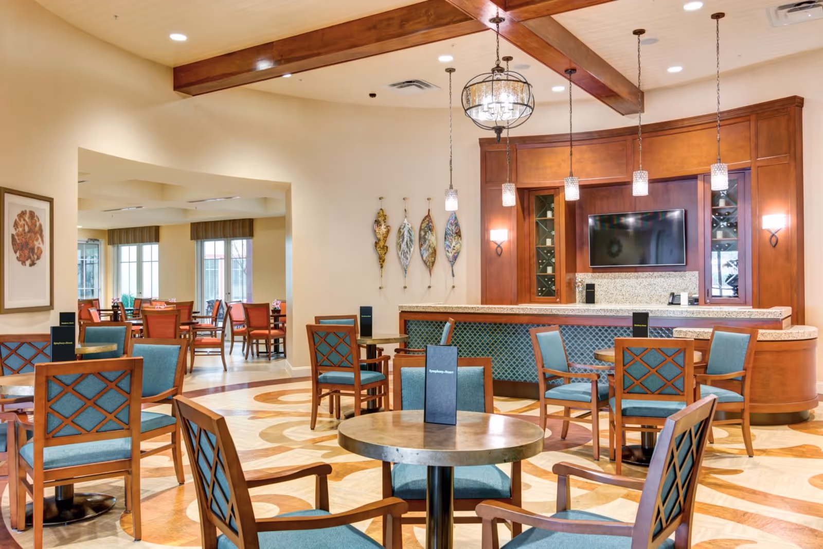 A spacious and well-lit dining area in a senior living facility with round tables and wooden chairs featuring blue cushions. The room has a decorative wooden ceiling beam, pendant lights, and a bar area with a mounted flat-screen TV. The floor has an intricate circular pattern, and there is wall art and large windows letting in natural light.