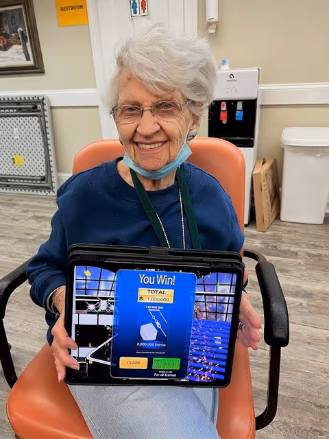 An elderly woman with white hair and glasses is sitting in an orange chair indoors, smiling and holding a tablet displaying a game screen that says 'You Win!' with a total prize of 1,000,000. She is wearing a blue sweater and has a face mask pulled down below her chin. Behind her, there is a water dispenser, a trash can, and a sign indicating a restroom.