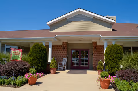 Brick front entrance with white columns, potted flowers, and glass double doors under a covered walkway.