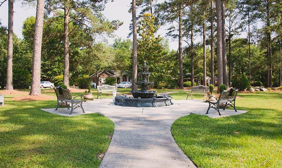 A peaceful outdoor garden area with a central stone fountain surrounded by four metal benches on a paved circular platform. Tall pine trees and green grass surround the area, with a house and parked cars visible in the background under a clear sky.