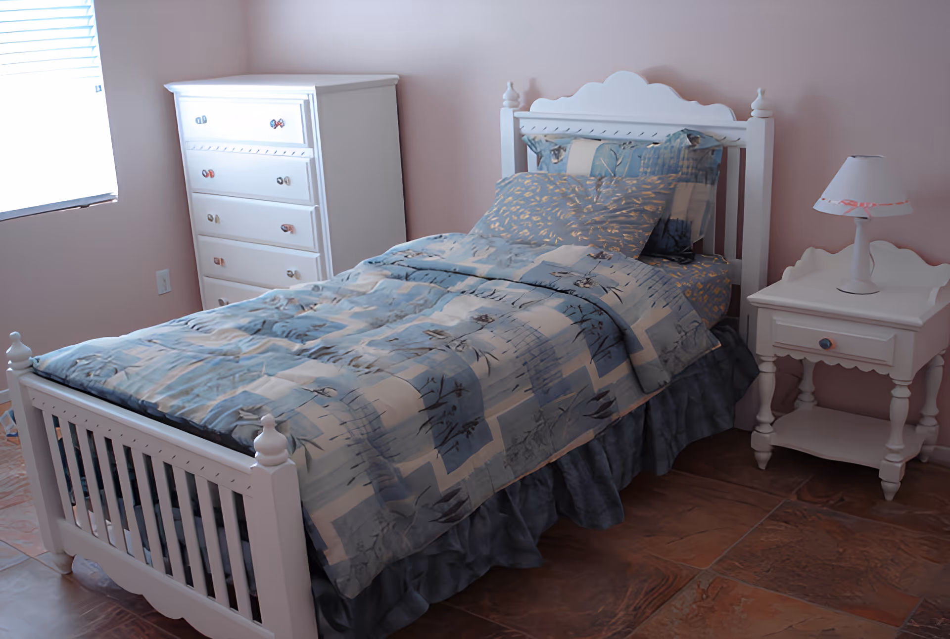 A bedroom with a white wooden bed frame, a bed made with blue and white patterned bedding, a white nightstand with a small lamp, and a white chest of drawers near a window with blinds.