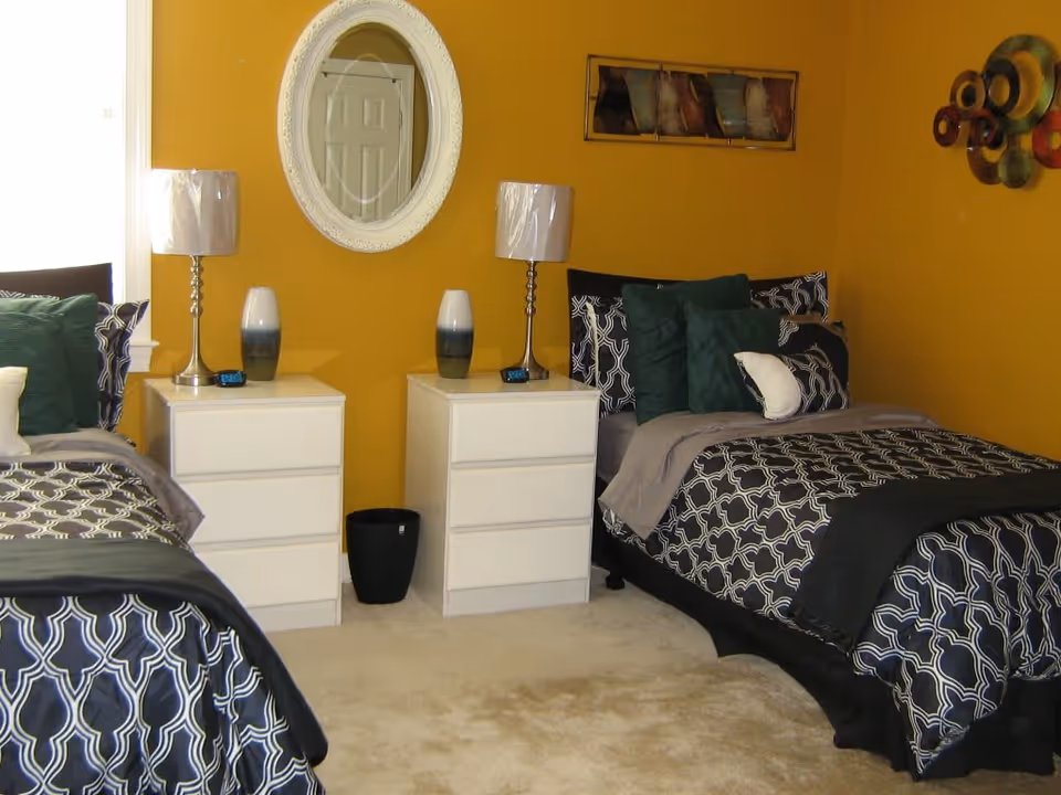 Two twin beds with patterned bedding in a mustard-yellow bedroom with white nightstands, lamps, and a round wall mirror.
