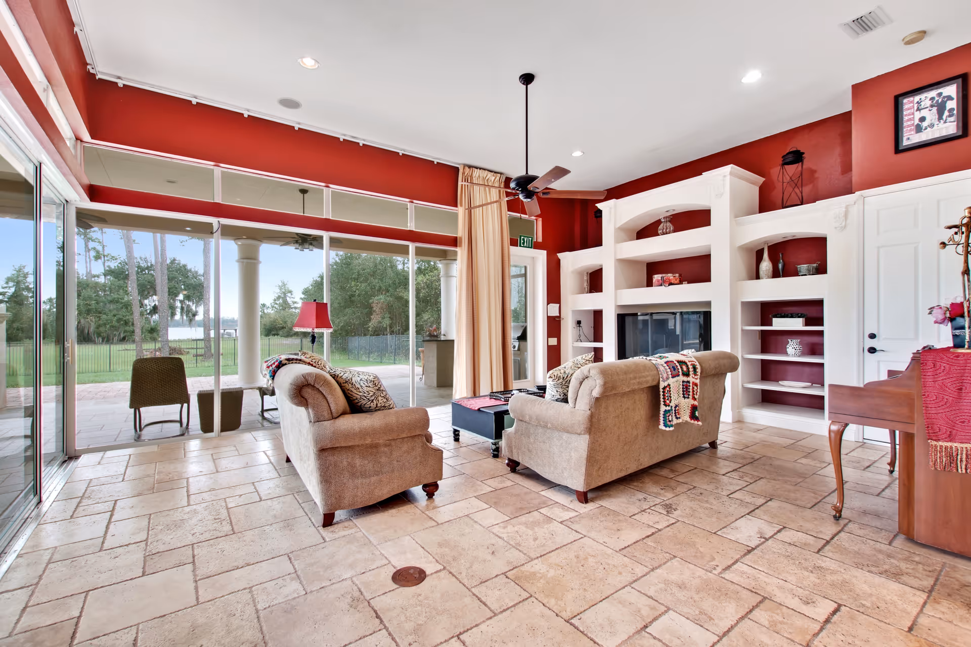 A bright living room with two beige upholstered sofas facing a white built-in shelving unit and fireplace. The room features large floor-to-ceiling glass sliding doors that open to an outdoor patio with a view of trees and a lake. The walls are painted red above the white shelves, and the floor is tiled with light-colored stone. A ceiling fan hangs above, and there is a piano with a red cloth on the right side of the room.