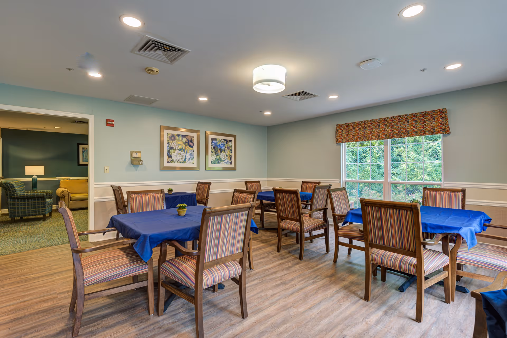 A dining room with several wooden tables covered with blue tablecloths and surrounded by wooden chairs with striped cushions. The room has light blue walls with white wainscoting, two framed floral paintings on the wall, a large window with a patterned valance, and a ceiling light fixture. Through an open doorway, a living area with armchairs and a lamp is visible.