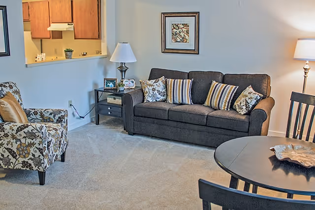 Cozy living room with a dark sofa and patterned armchair, side tables and lamps, a small round dining table, and a pass-through to the kitchen.