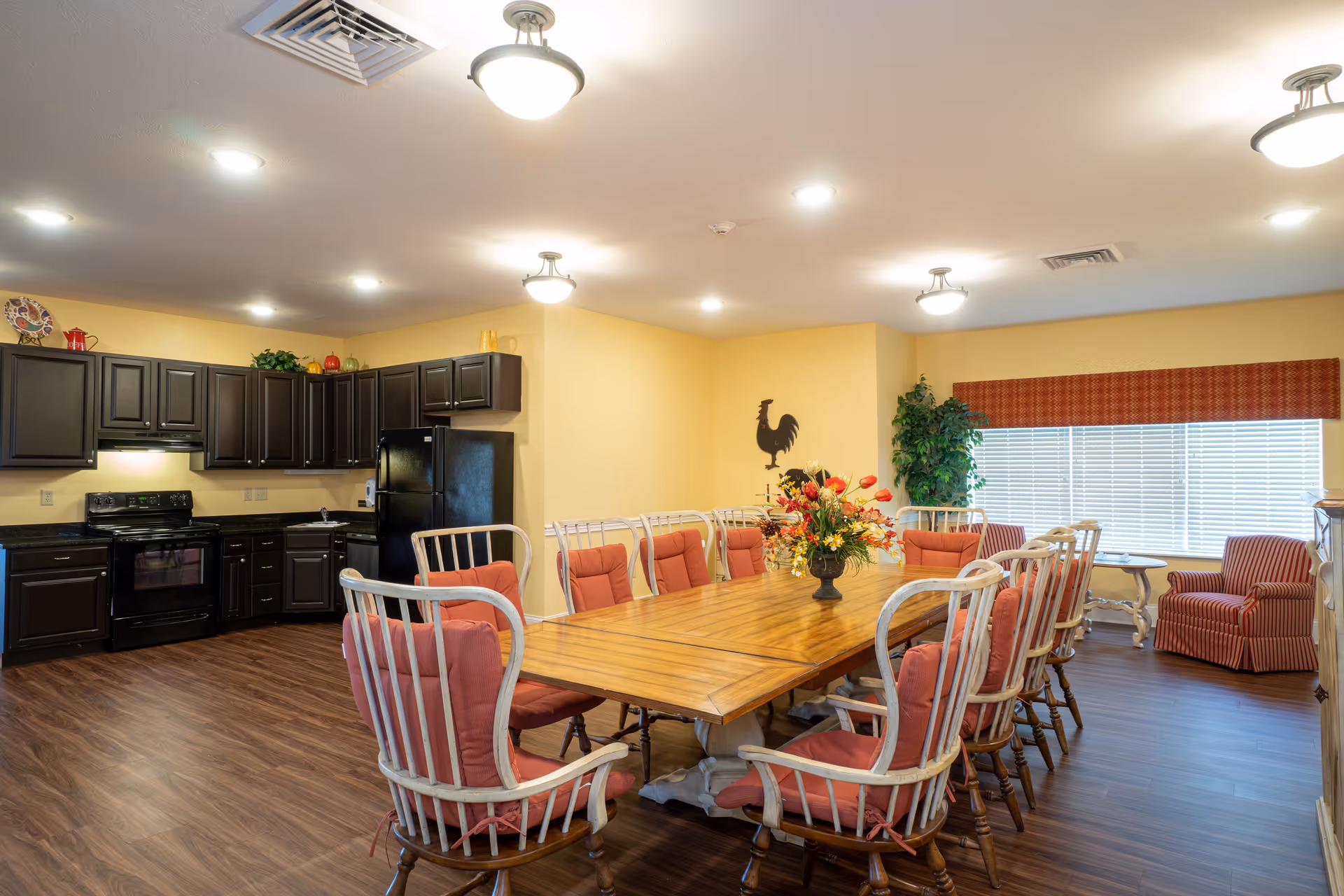 A spacious dining area with a large wooden table surrounded by cushioned chairs with red upholstery. The room features dark wood kitchen cabinets, a black refrigerator, stove, and sink along one wall. The floor is a dark wood laminate, and the walls are painted a soft yellow. There is a window with blinds and a red valance, a decorative rooster wall hanging, a floral centerpiece on the table, and a potted plant in the corner.