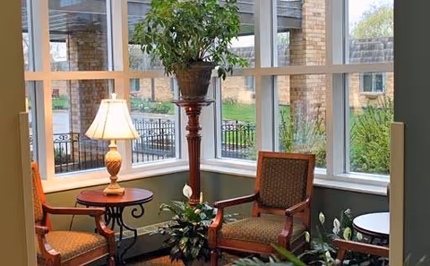 A cozy sitting area inside Friendship Manor featuring two upholstered armchairs with wooden frames, a small round wooden table with a lamp, a tall plant stand with a potted plant, and large windows providing a view of the outdoor garden area.