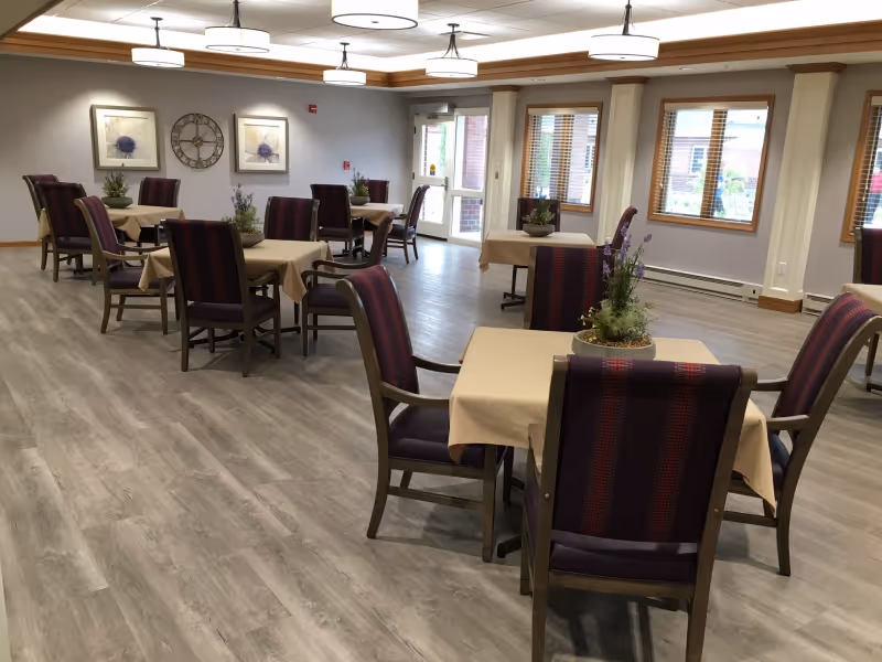 A spacious dining room with several square tables covered with beige tablecloths, each surrounded by four chairs with dark upholstery and red stripes. The room has light wood flooring, large windows with blinds, and ceiling lights. There are decorative plants on each table, a large clock, and framed artwork on the wall.