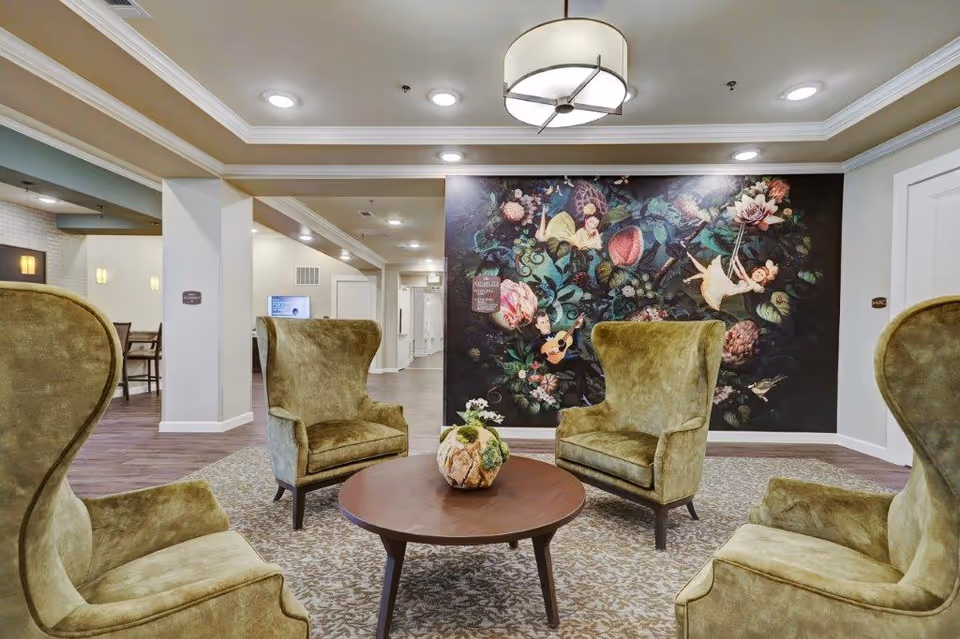 A lounge area with four green wingback chairs arranged around a round wooden coffee table in front of a large floral mural.