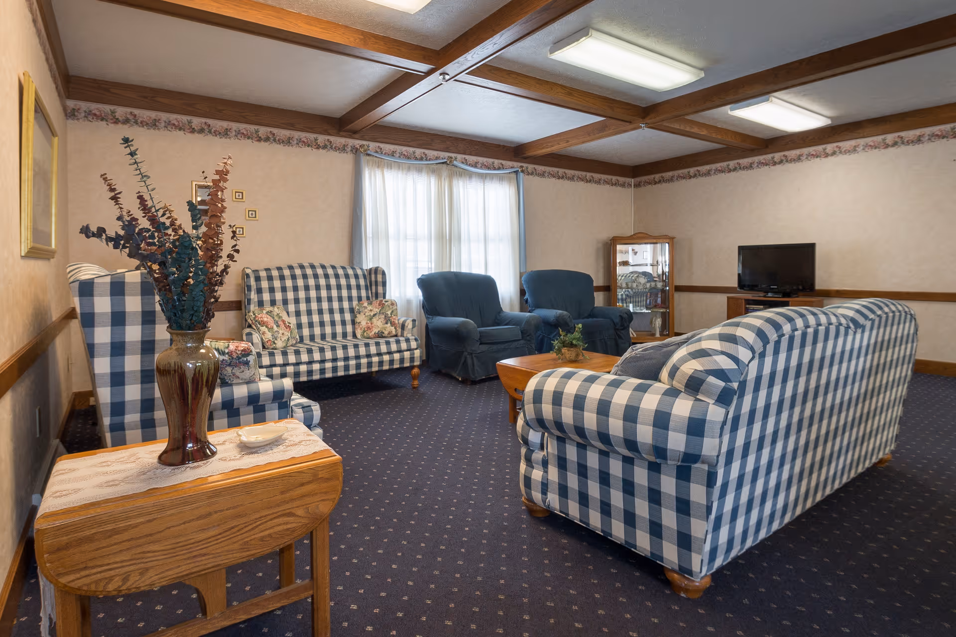 A cozy living room with blue and white checkered sofas and armchairs, two solid blue armchairs, a wooden coffee table with a small plant, a wooden side table with a vase of dried flowers, a TV on a wooden stand, and a window with sheer curtains. The room has a patterned carpet, floral wallpaper border, and wooden ceiling beams.