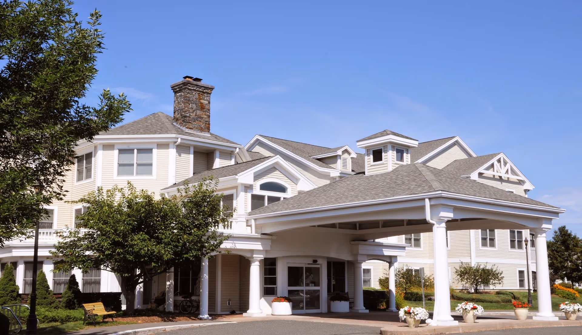 The front façade and covered porte-cochère of a multi-story retirement community building with columns, landscaping, and a clear blue sky.