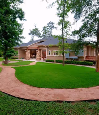 Exterior view of a single-story building with a covered entrance, surrounded by green grass, trees, and a curved walkway in front.