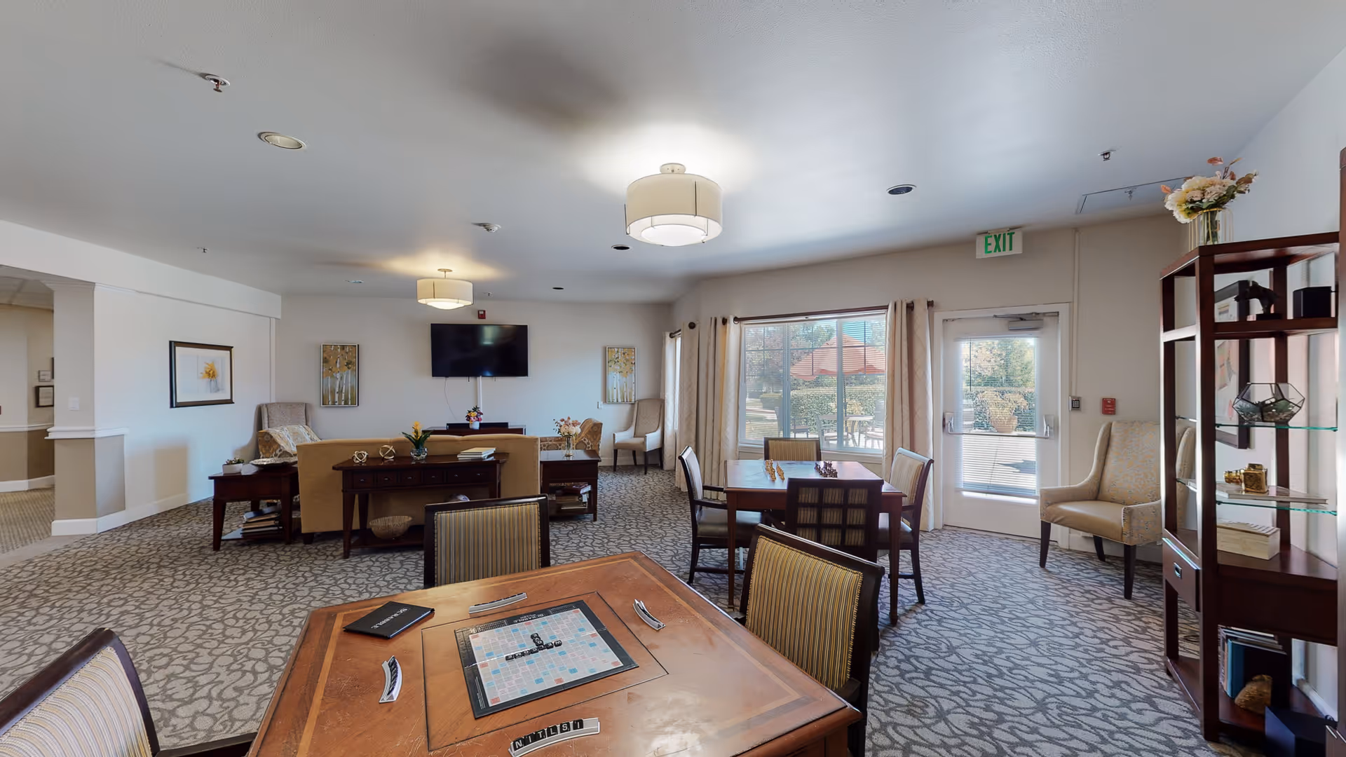 A spacious senior living common area with a patterned carpet floor, several tables and chairs including a game table with a Scrabble board, a seating area with sofas and armchairs, a wall-mounted TV, large windows with curtains letting in natural light, and a glass door leading outside. There is a wooden shelving unit with decorative items and flowers on top.