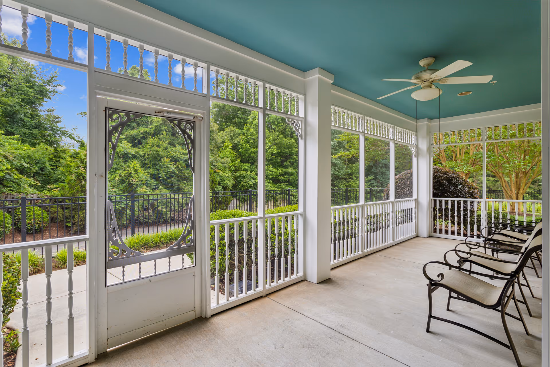 A screened-in porch with white railings and decorative trim, featuring four metal chairs with cushions lined up along the right side. The porch has a teal ceiling with a white ceiling fan. Outside the porch, there is lush green landscaping with trees and bushes under a blue sky with some clouds.