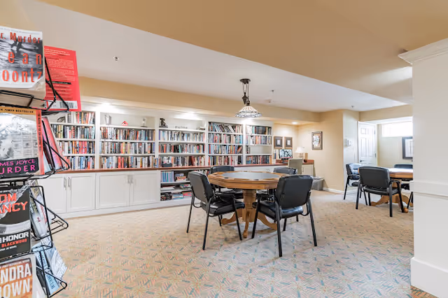 Community library/reading room with built-in bookshelves, a round table surrounded by chairs, and a magazine rack.