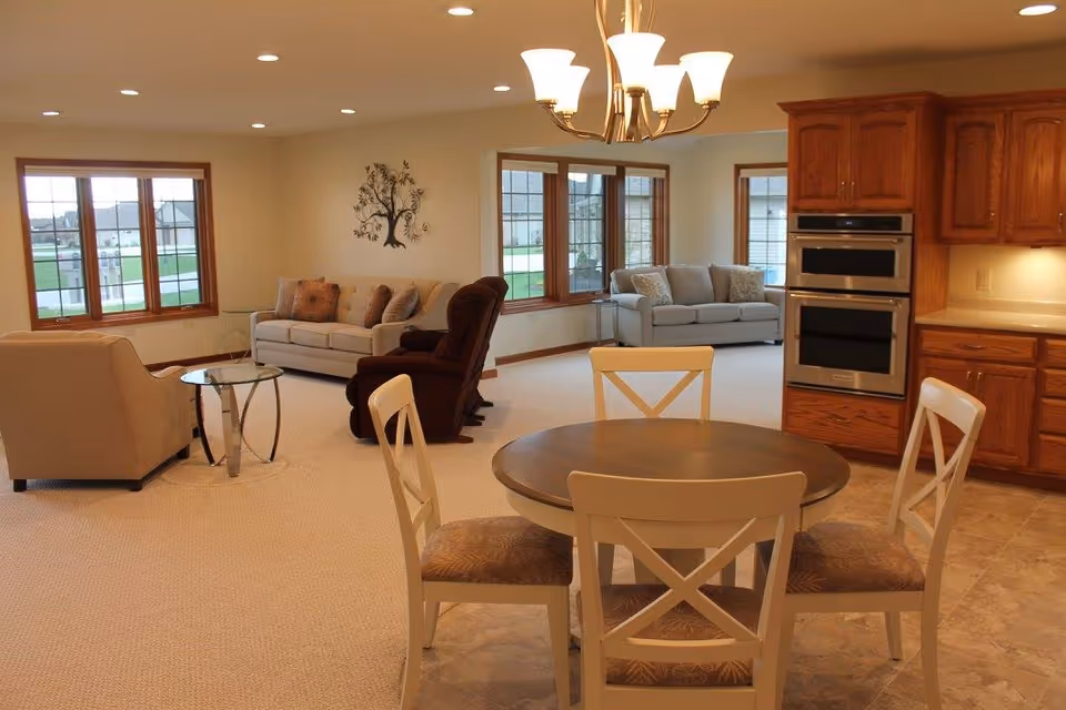 Open-plan interior with a round dining table and chairs in the foreground, a living room with sofas and recliner near large windows, and a kitchen area with wooden cabinets in the background.