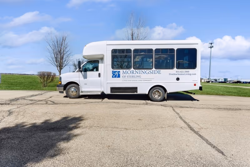 A white shuttle bus parked on a paved area with grass and trees in the background under a blue sky with some clouds. The bus has the logo and text 'Morningside of Sterling' along with contact information and the website FiveStarSeniorLiving.com on its side.