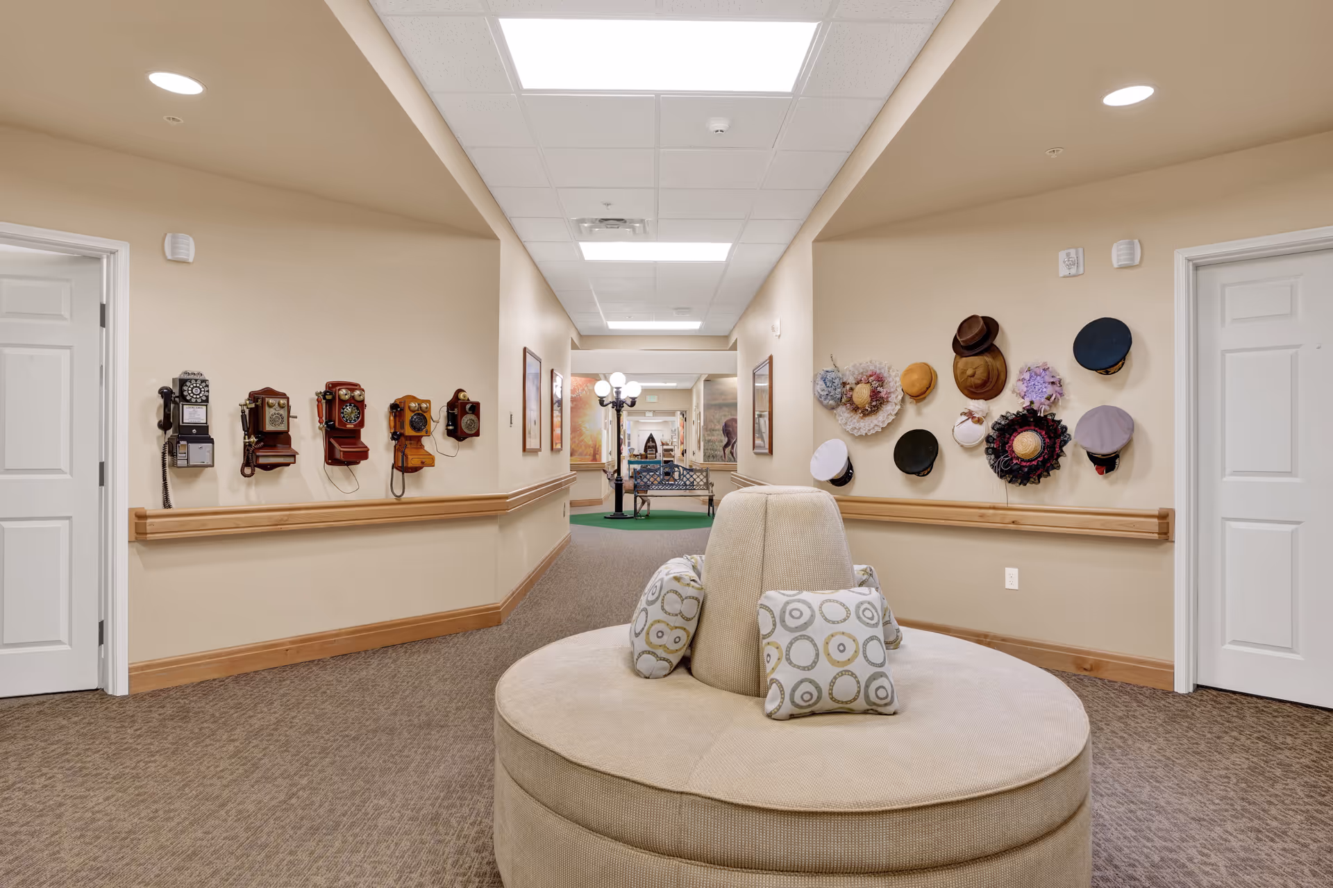A hallway in a senior living facility with beige walls and carpeted floor. On the left wall, there are several vintage rotary telephones mounted. On the right wall, various hats are displayed as decoration. In the center of the hallway is a round beige cushioned bench with patterned pillows. The hallway extends to a common area with a bench and lamp post visible in the background.