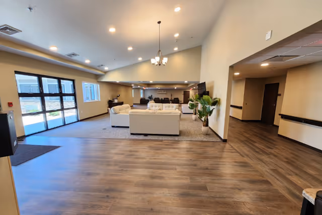 Spacious senior living common area with sofas, potted plants, large windows, and wood-look flooring.