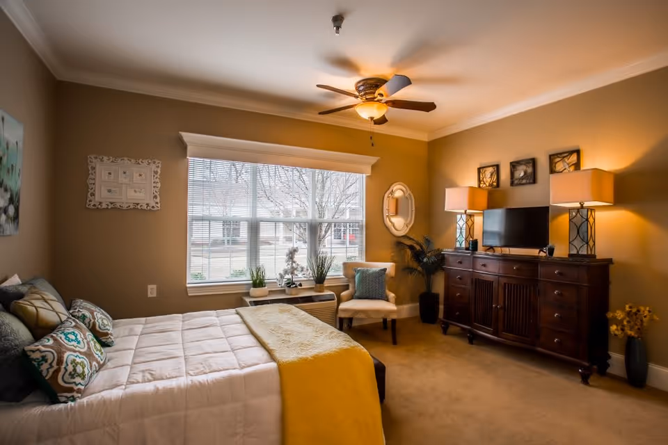 A cozy bedroom in a senior living facility featuring a neatly made bed with decorative pillows and a yellow throw blanket. The room has a large window with blinds, a comfortable armchair with a cushion, a wooden dresser with a flat-screen TV, two table lamps, and wall decorations. The walls are painted beige, and the ceiling has a fan with a light fixture.