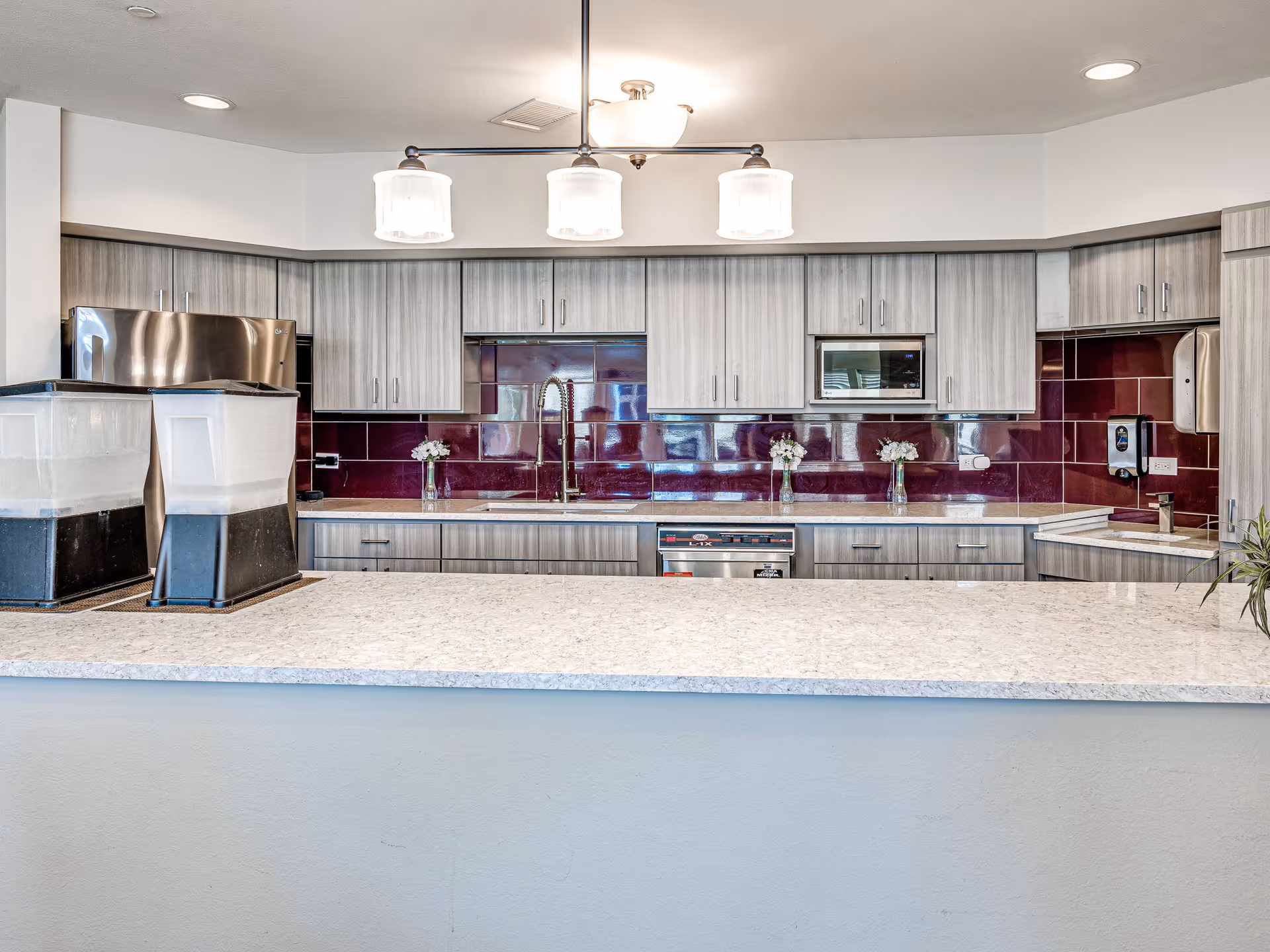 Modern kitchen area with light gray wooden cabinets, a stainless steel refrigerator, microwave, and dishwasher. The backsplash features glossy dark red tiles. There are two beverage dispensers on the light-colored countertop, and three small vases with white flowers are placed along the counter near the sink. A ceiling light fixture with three frosted glass shades hangs above.