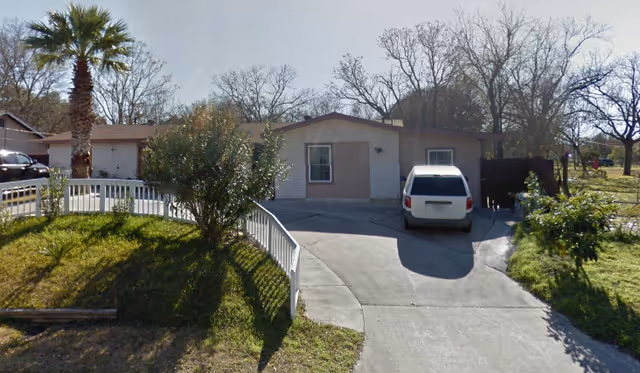 Single-story residential building with a driveway and a white vehicle parked in front. The building has a garage door and windows with shutters. There are trees and bushes around the property, and a white fence along the driveway.