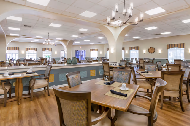 A spacious dining room with multiple wooden tables and chairs arranged neatly. The room features arched openings, large windows with striped valances, and a wooden floor. There are chandeliers and wall sconces providing lighting, and a clock is mounted on one wall.