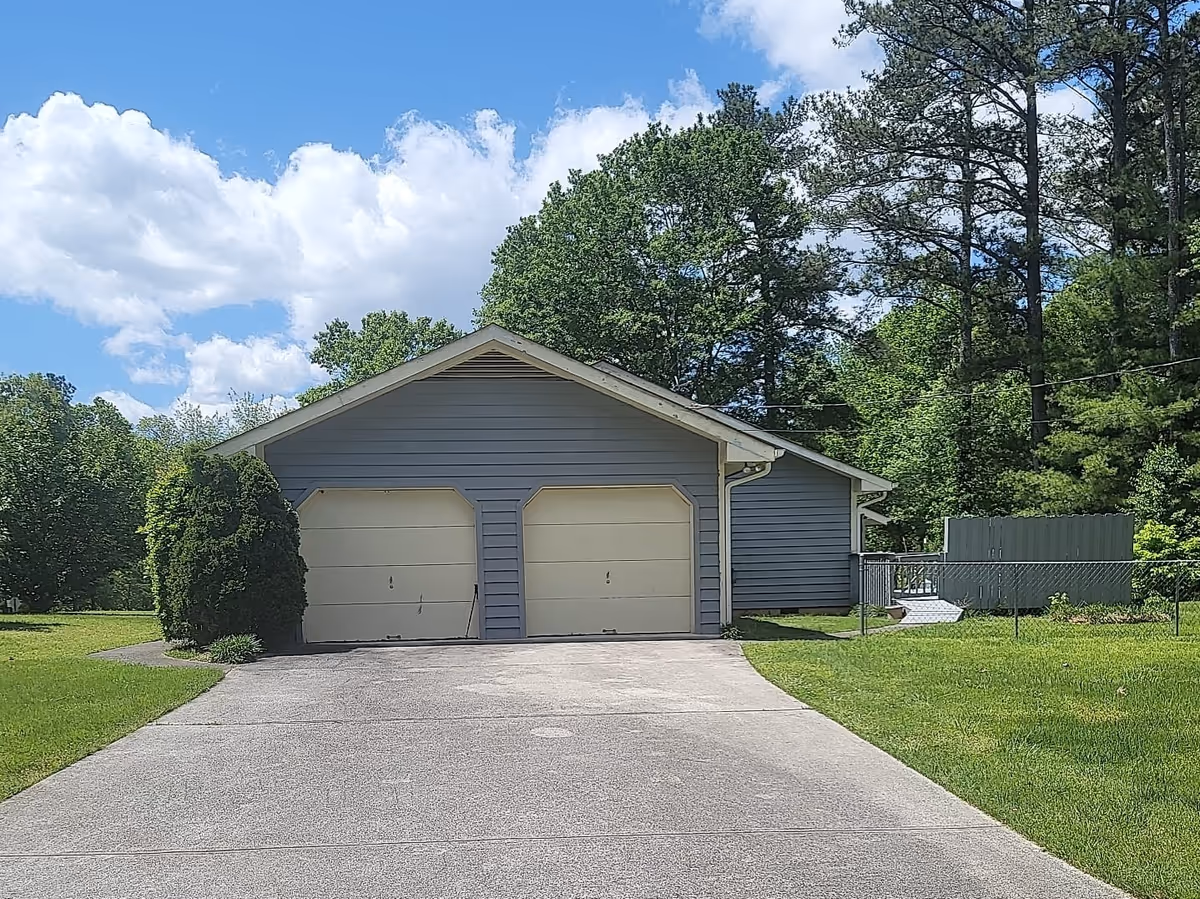 A two-car garage with closed beige doors at the end of a concrete driveway, surrounded by lawn and trees under a blue sky.
