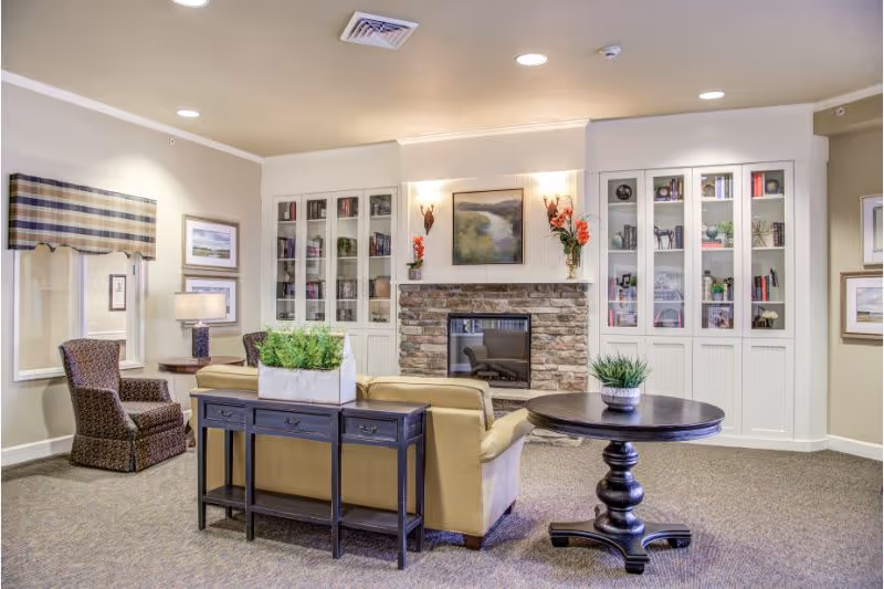 A cozy living room area in a senior living facility featuring a stone fireplace with a painting above it, built-in white cabinets with glass doors filled with books and decor, a beige sofa, a dark wooden console table with a plant, a round wooden table with a small plant, and two patterned armchairs near a window with a plaid valance.
