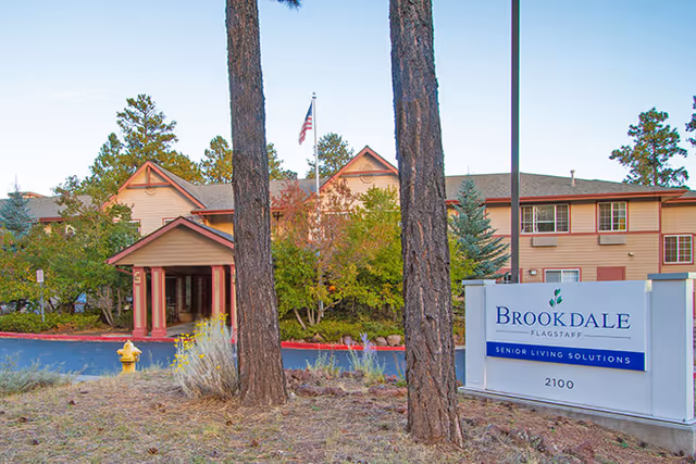 Exterior view of the Brookdale Flagstaff senior living facility building with trees and a sign in the foreground that reads 'Brookdale Flagstaff Senior Living Solutions 2100'.
